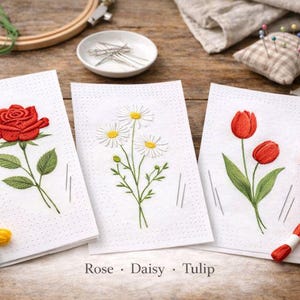 May include: Three embroidered floral designs on white fabric: a red rose, white daisies, and red tulips. The text "Rose • Daisy • Tulip" is below the designs. Embroidery floss and a wooden hoop are also visible.