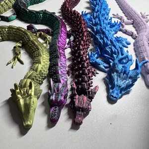 Peut inclure: Plusieurs figurines de dragons articulés aux couleurs variées, vert, violet, bleu et rose. Les dragons, avec leurs écailles détaillées, sont disposés sur une surface blanche. Ces objets décoratifs sont probablement imprimés en 3D.
