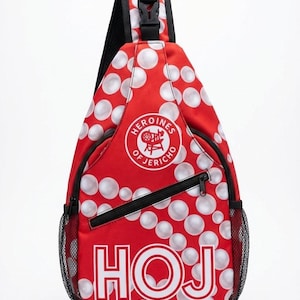 Puede incluir: Mochila bandolera roja con lunares blancos y el texto "HOJ". La bolsa tiene un bolsillo lateral de malla negra, una cremallera negra y una correa negra. También tiene un logotipo circular que dice "Heroines of Jericho".
