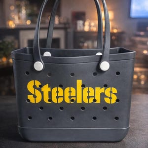May include: A dark gray tote bag with black handles and the word "Steelers" in yellow stencil font. The bag has small circular holes and white fasteners. Perfect for carrying items.