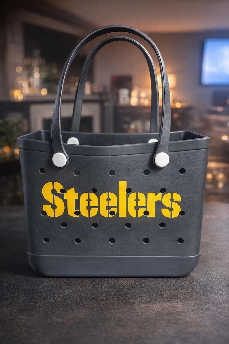 May include: A dark gray tote bag with black handles and the word "Steelers" in yellow stencil font. The bag has small circular holes and white fasteners. Perfect for carrying items.
