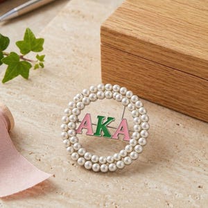Puede incluir: Broche circular con borde de perlas, con las letras AKA en rosa y verde. El broche tiene un alfiler. En el fondo hay una caja de madera, un bolígrafo y una ramita de hiedra.