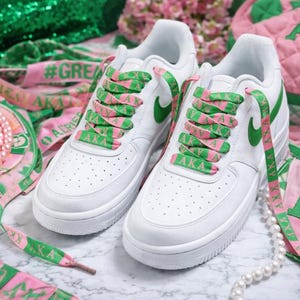 Może przedstawiać: Białe trampki z zielonymi i różowymi akcentami, w tym sznurowadłami i logo Nike. Sznurowadła mają litery "AKA". Akcesoria obejmują naszyjnik z pereł oraz różowe i zielone wstążki.