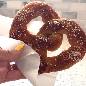 May include: A soft pretzel, golden brown with coarse salt, is held in a white paper wrapper. The pretzel is a classic baked snack, perfect for a quick bite. The background is a light-colored countertop and a tiled wall.