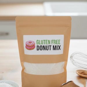 May include: A brown paper bag of gluten-free donut mix. The label features a pink donut illustration and the words "GLUTEN FREE DONUT MIX" in green and black. A bowl of white powder and a whisk are in the background.