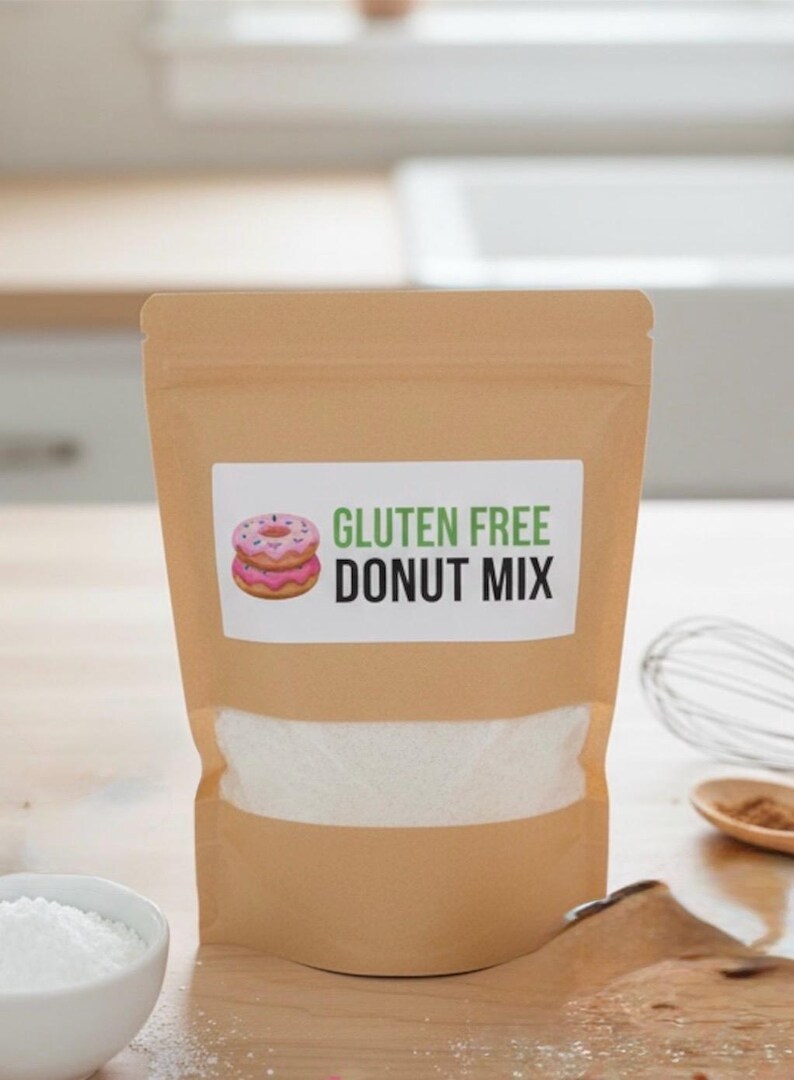 May include: A brown paper bag of gluten-free donut mix. The label features a pink donut illustration and the words "GLUTEN FREE DONUT MIX" in green and black. A bowl of white powder and a whisk are in the background.