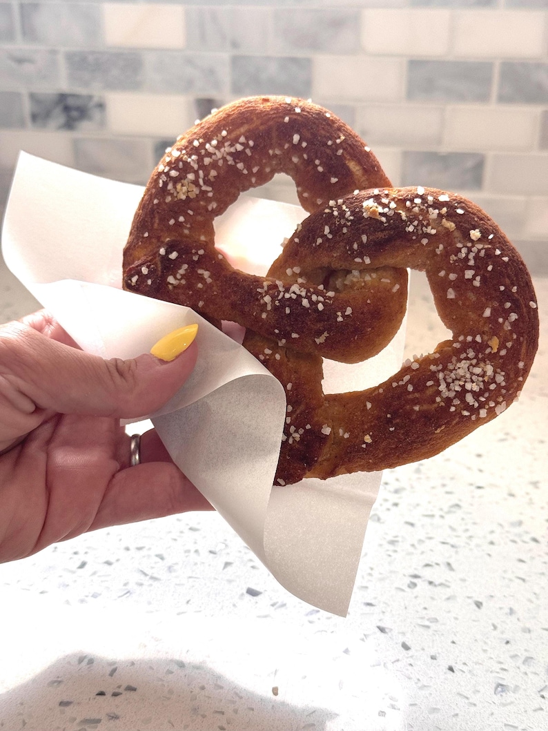 May include: A soft pretzel, golden brown with coarse salt, is held in a white paper wrapper. The pretzel is a classic baked snack, perfect for a quick bite. The background is a light-colored countertop and a tiled wall.
