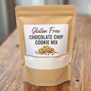 May include: A brown paper bag of gluten-free chocolate chip cookie mix. The bag has a white label with the words "Gluten Free Chocolate Chip Cookie Mix" and an illustration of three cookies with chocolate chips. The bag is partially filled with white powder.