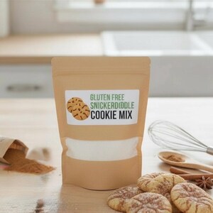 May include: A tan bag of gluten-free snickerdoodle cookie mix with a white label featuring cookie illustrations. Several baked cookies, cinnamon, and a whisk are arranged on a wooden surface, suggesting a baking theme.