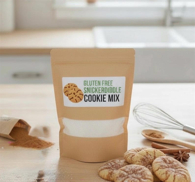 May include: A tan bag of gluten-free snickerdoodle cookie mix with a white label featuring cookie illustrations. Several baked cookies, cinnamon, and a whisk are arranged on a wooden surface, suggesting a baking theme.