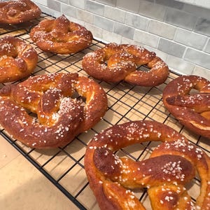 May include: Several freshly baked pretzels with a golden-brown crust and sprinkled with coarse salt are arranged on a black wire cooling rack. The pretzels are a classic baked good, perfect for a snack or appetizer.