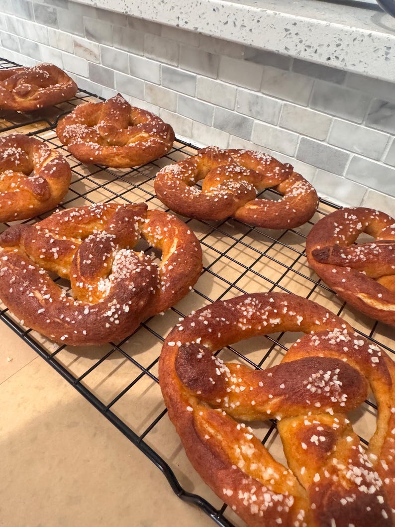 May include: Several freshly baked pretzels with a golden-brown crust and sprinkled with coarse salt are arranged on a black wire cooling rack. The pretzels are a classic baked good, perfect for a snack or appetizer.