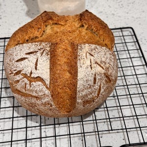 May include: A freshly baked loaf of bread with a golden-brown crust, resting on a black wire rack. The bread has decorative cuts on the surface and is dusted with flour, showcasing a rustic, artisanal appearance. The bread is a round shape.
