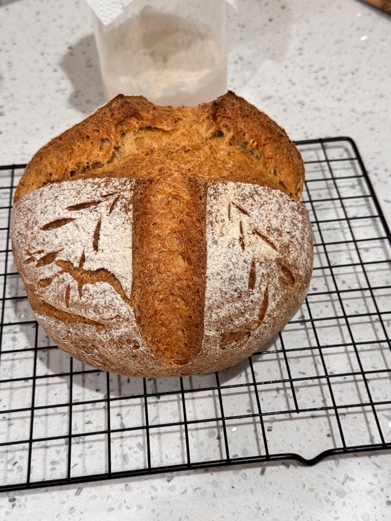 May include: A freshly baked loaf of bread with a golden-brown crust, resting on a black wire rack. The bread has decorative cuts on the surface and is dusted with flour, showcasing a rustic, artisanal appearance. The bread is a round shape.
