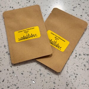 May include: Two brown paper packets with yellow labels. The labels read "Gluten Free Dehydrated Starter" with a graphic of plants. The packets are on a speckled, light gray surface.