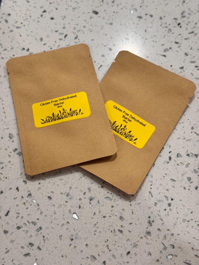 May include: Two brown paper packets with yellow labels. The labels read "Gluten Free Dehydrated Starter" with a graphic of plants. The packets are on a speckled, light gray surface.