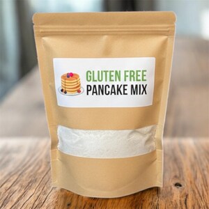 May include: A brown paper bag of gluten-free pancake mix. The bag has a clear window showing the white mix. A white label displays the text "GLUTEN FREE PANCAKE MIX" with a stack of pancakes illustration.