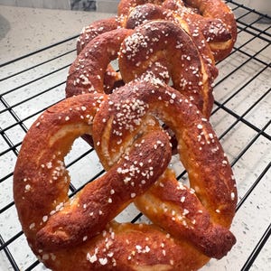 May include: Several freshly baked pretzels with a golden-brown crust and sprinkled with coarse salt are arranged on a black wire rack. The pretzels have a classic knotted shape and are ready to eat.