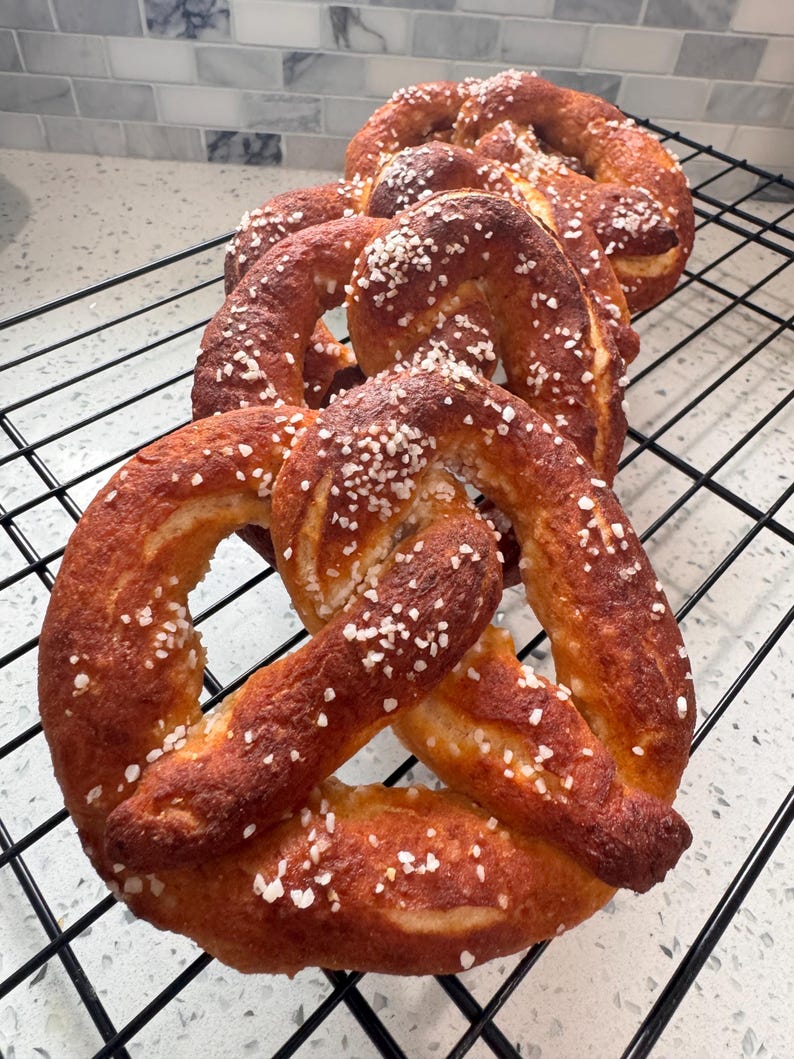 May include: Several freshly baked pretzels with a golden-brown crust and sprinkled with coarse salt are arranged on a black wire rack. The pretzels have a classic knotted shape and are ready to eat.