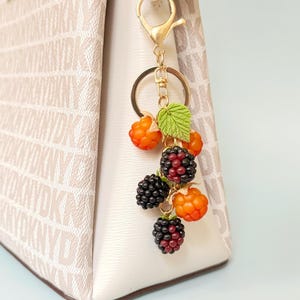 May include: A gold-toned keychain charm featuring a cluster of miniature, detailed berries. The charm includes black and red blackberries, orange raspberries, and a green leaf. The charm is attached to a beige handbag.