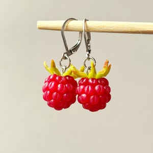 May include: A pair of dangle earrings featuring bright red raspberry charms with green and yellow accents. The earrings are suspended from silver-colored lever-back ear wires, showcasing a playful, fruit-themed design.
