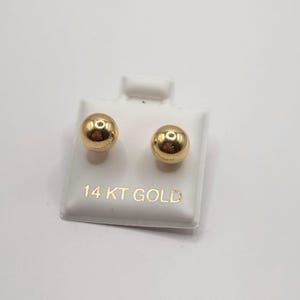 May include: A pair of polished, round, gold-colored stud earrings. The earrings are displayed on a white card with the text "14 KT GOLD" in gold lettering.