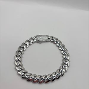 May include: A silver-colored curb chain bracelet with a rectangular clasp. The bracelet is made of interlocking links and has a polished finish. The clasp is encrusted with small, clear stones.