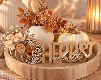 May include: A round wooden tray containing a white vase with dried orange flowers, a white pumpkin with string lights, and wooden letters spelling HERBST. A cutout pumpkin shape features the text HALLO HERBST and the word verliebt with a heart.