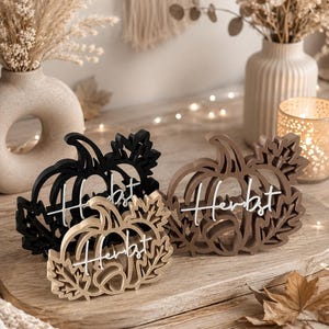 Può includere: Tre ornamenti decorativi a forma di zucca in nero, marrone e beige, ognuno caratterizzato da intricati ritagli di foglie e dalla parola "Herbst" in caratteri bianchi al centro. Questi articoli sono disposti insieme come un set.