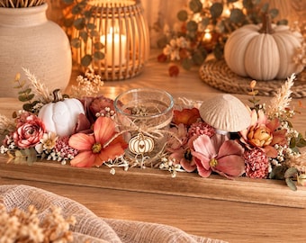 May include: A rectangular wooden tray centerpiece featuring a clear glass candle holder with a small wooden pumpkin charm, surrounded by artificial flowers in shades of pink and orange, a white ceramic pumpkin, and a beige mushroom figurine.