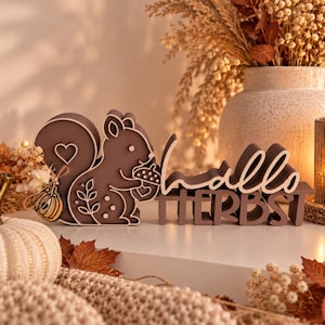 Op de afbeelding: Een bruin houten eekhoornfiguur dat een eikel vasthoudt, met een hartvormige uitsparing in de staart en een kleine decoratieve pompoenbedel. Daarnaast staat een bruin houten bord met de tekst "hallo HERBST" in een mix van schrijf- en blokletters.
