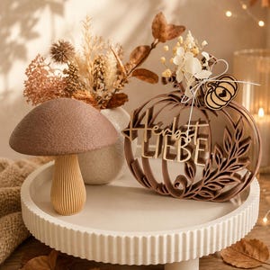 Könnte beinhalten: Eine braune Pilzfigur mit strukturiertem Hut und geripptem Stiel neben einer braunen, lasergeschnittenen Holzkürbis-Dekoration mit dem Schriftzug Herbst LIEBE, verziert mit einem kleinen Holzkürbis-Anhänger und getrockneten weißen Blumen.