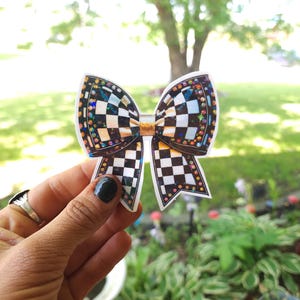 May include: A decorative bow sticker with a black and white checkered pattern. The bow has a gold center and is outlined with colorful rhinestones. The sticker is held up against a blurred background of green foliage and a tree.