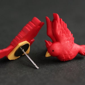 Puede incluir: Un par de pendientes de p&aacute;jaro cardenal rojo. Los pendientes est&aacute;n hechos de arcilla polim&eacute;rica y tienen un dise&ntilde;o realista. Los pendientes est&aacute;n unidos a postes de plata.
