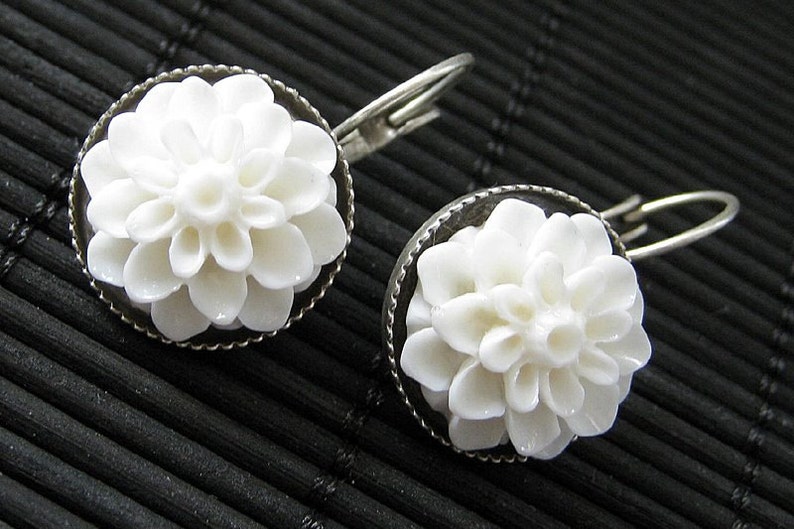 May include: A pair of white flower earrings with silver hooks. The flowers are made of a white material that looks like porcelain or ceramic. The earrings are a delicate and feminine accessory.