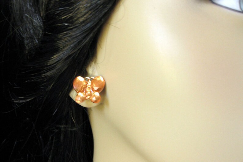 May include: A pair of copper butterfly earrings. The earrings are a light orange color and have a textured surface.