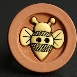 May include: A brown button with a yellow bee design. The bee has black eyes and a black and yellow striped body.