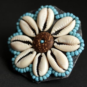 May include: A white shell flower with a brown center, surrounded by blue beads on a black background.
