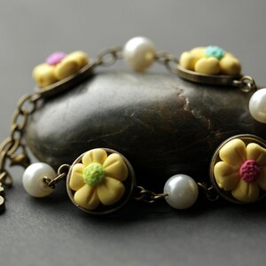 May include: A yellow flower charm bracelet with white pearl beads and a bronze chain. The bracelet has a letter 'S' charm.
