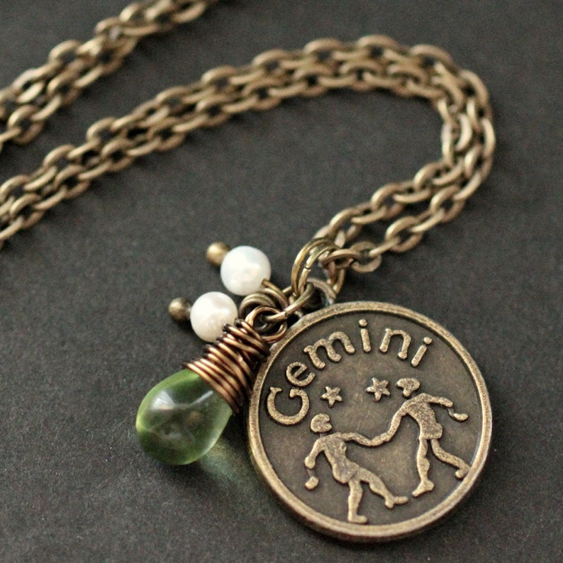 May include: A gold necklace with a bronze pendant featuring the Gemini zodiac sign and the word "Gemini". The pendant is adorned with a green glass bead and three white pearl beads.
