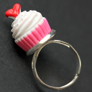 May include: A silver ring with a pink and white cupcake charm. The cupcake has a red heart on top.
