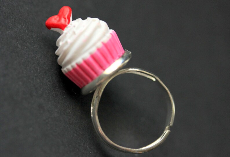 May include: A silver ring with a pink and white cupcake charm. The cupcake has a red heart on top.