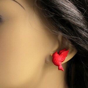 Puede incluir: Pendientes de bot&oacute;n de p&aacute;jaro rojo. Los pendientes est&aacute;n hechos de un material pl&aacute;stico rojo y tienen forma de p&aacute;jaro en vuelo. Los pendientes se muestran en la oreja de un maniqu&iacute;.