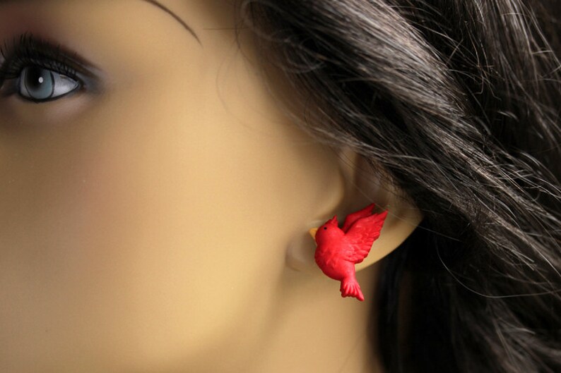 Puede incluir: Pendientes de bot&oacute;n de p&aacute;jaro rojo. Los pendientes est&aacute;n hechos de un material pl&aacute;stico rojo y tienen forma de p&aacute;jaro en vuelo. Los pendientes se muestran en la oreja de un maniqu&iacute;.