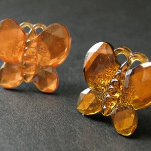 May include: A pair of amber-colored butterfly-shaped earrings. The earrings are made of faceted glass and have a shiny finish.