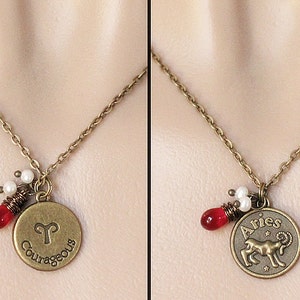 Może przedstawiać: Złoty naszyjnik z zawieszką przedstawiającą astrologiczny symbol Barana, barana. Zawieszka zawiera r&oacute;wnież napis "Aries" i "Odważny". Naszyjnik ma czerwone i białe koraliki.