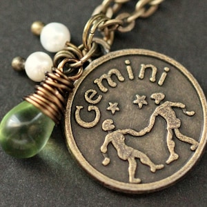 May include: A bronze pendant necklace with a Gemini zodiac sign charm, two white pearl beads, and a green glass teardrop charm.