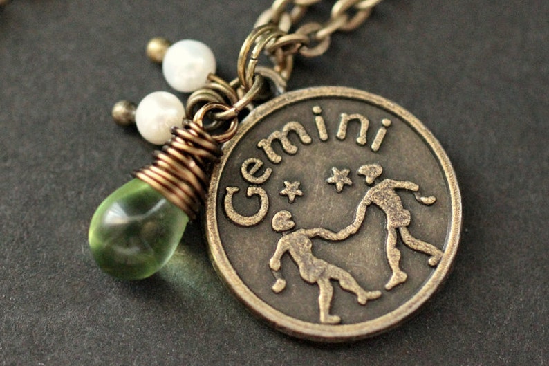 May include: A bronze pendant necklace with a Gemini zodiac sign charm, two white pearl beads, and a green glass teardrop charm.