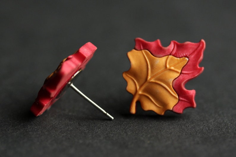 Pu&ograve; includere: Un paio di orecchini a forma di foglia rossa e arancione con perni d'argento. Gli orecchini sono realizzati in pasta polimerica e hanno una superficie testurizzata.
