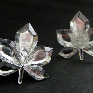 May include: A pair of clear crystal earrings shaped like maple leaves. The leaves have a faceted design and are set on a silver tone metal.